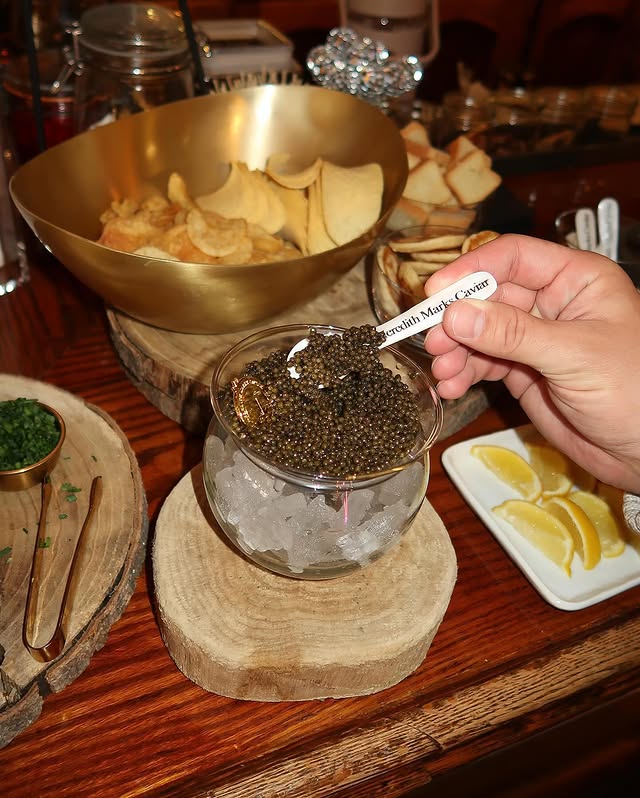 Bowl of Meredith Marks Caviar on ice served with a mother-of-pearl spoon, chips, and lemon wedges on a dinner table.