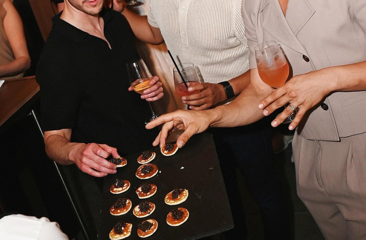 Alt text: Guests enjoying blinis topped with caviar and champagne cocktails at an elegant gathering, highlighting a refined caviar and champagne pairing experience.